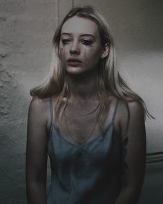 a woman with long blonde hair standing in front of a white wall and looking at the camera