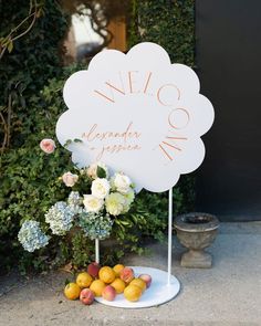 a white sign sitting on top of a table next to flowers and fruit in front of a bush