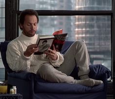 a man sitting on a blue couch reading a book in front of a large window