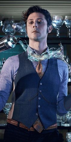 a man in a vest and tie standing next to a shelf filled with wine glasses