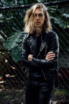 a man with long hair standing in front of a fence wearing a black leather jacket