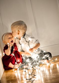 two toddlers sitting on the floor with christmas lights around them and one is kissing the other's cheek