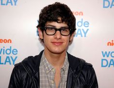 a young man wearing glasses standing in front of a wall