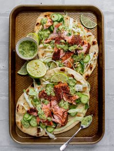 two tacos with meat, vegetables and sauces on a baking tray next to a spoon