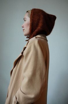 a woman wearing a coat with a hat on it's head and standing in front of a wall