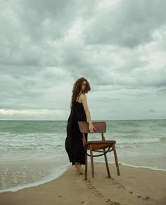 a woman standing on top of a wooden chair near the ocean under a cloudy sky