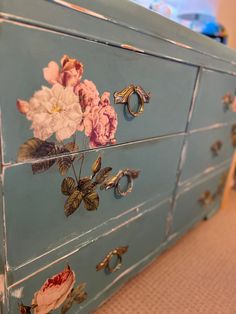 a blue dresser with flowers painted on the drawers and handles, sitting on carpeted flooring