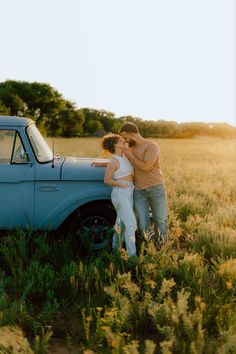 a man and woman standing next to an old truck in a field with tall grass