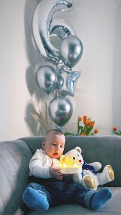 a baby is sitting on a couch with a teddy bear and some balloons in the background