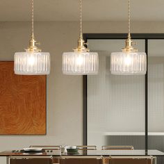 three glass pendant lights hanging over a dining room table with chairs and plates on it