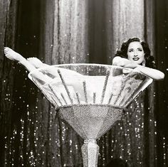 a black and white photo of a woman sitting in a martini glass with her legs spread out