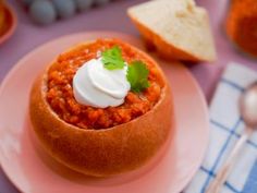 a bowl of chili with sour cream on top sits on a pink plate next to bread and silverware