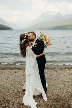 a bride and groom kissing on the shore of a lake with mountains in the background