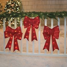 three red bows are hanging on a fence with christmas lights in the snow behind them