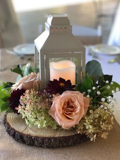 a small white lantern with flowers and greenery on it sitting on a wooden slice