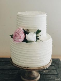 a three layer white cake with pink and white flowers on top, sitting on a wooden table