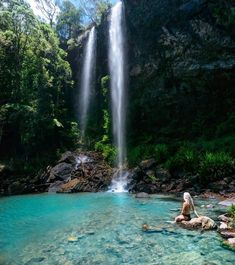 a woman sitting on rocks in front of a waterfall with blue water and green trees