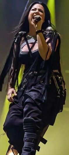 a woman with long black hair is singing into a microphone and holding her hands in the air