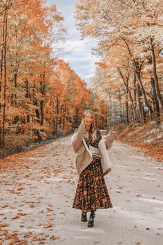 a woman standing in the middle of an empty road surrounded by trees with orange leaves