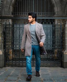 a man walking down the street in front of an iron gate wearing a gray coat and blue jeans