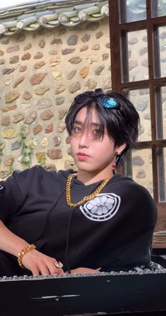 a young man sitting in front of a window wearing a black shirt and gold necklace