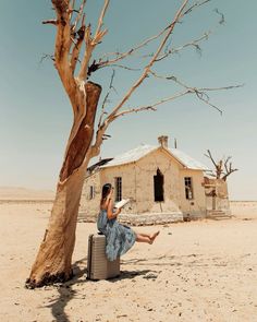 a woman sitting on top of a suitcase next to a tree in the middle of nowhere