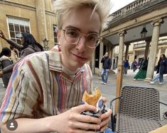 a young man sitting at an outdoor table with food in front of him and people walking around