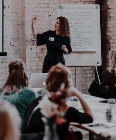 a woman standing in front of a whiteboard giving a presentation to people sitting at tables