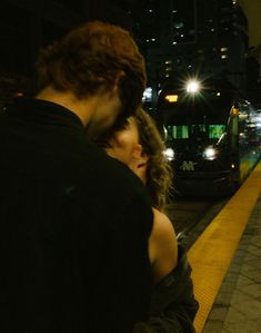 a man and woman standing next to each other on a sidewalk at night with city lights in the background