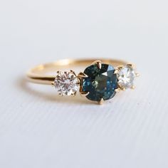 three stone engagement ring in yellow gold with blue and white diamonds on the side, sitting on a plain surface