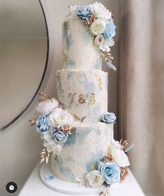 a three tiered wedding cake with blue and white flowers on the top, sitting on a table in front of a mirror