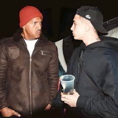 two men standing next to each other with one holding a cup and the other wearing a beanie