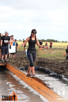 a woman riding a skateboard on top of a wooden beam in the mud while others watch