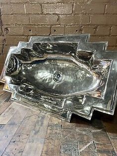 a metal fish shaped sink sitting on top of a wooden floor next to a brick wall