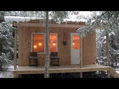 a small cabin in the woods with snow on the ground and two chairs sitting outside