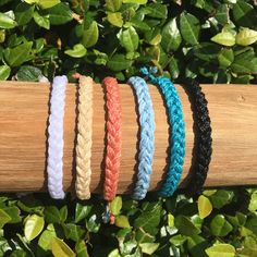 five braided bracelets sitting on top of a wooden stick next to green leaves