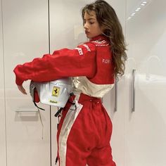 a woman in red and white racing suit leaning against a wall with a helmet on her shoulder