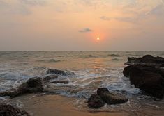 the sun is setting over the ocean with rocks in the foreground and waves on the shore