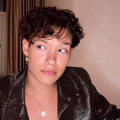 a woman with curly hair wearing a black jacket and gold necklace looking at the camera