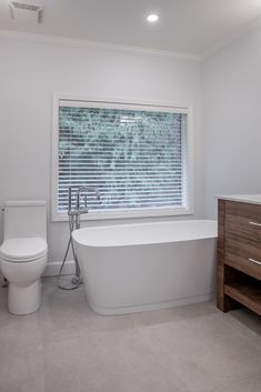 a white bath tub sitting next to a toilet in a bathroom under a window with blinds