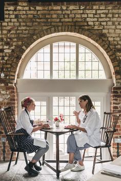 two women sitting at a table talking to each other in front of an arched window