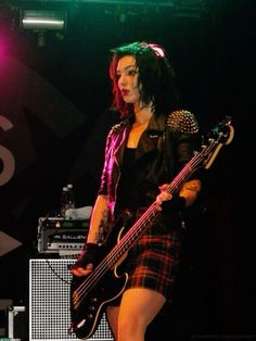 a woman with green hair is holding a bass in her hand and wearing a plaid skirt