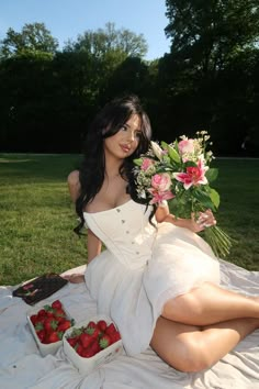 a woman in a white dress sitting on a blanket with strawberries and flowers