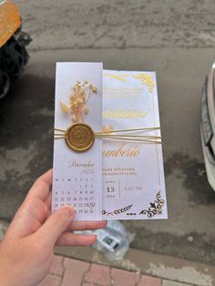 a person holding up a white and gold wedding card in front of a parked car