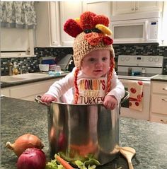 a baby in a kitchen with a pot and vegetables