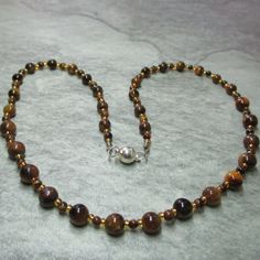 Tiger's Eye gemstone beads in shades of brown and three sizes make up this lovely necklace. Small Tiger's Eye beads run down the length of the necklace at each end they alternate with ten 6mm gemstone beads, while in the centre of the necklace the small Tiger's Eye's alternate with fourteen 8mm beads. Between all the Tiger's eye beads are silver lined gold coloured Glass seed beads. All the metal work including the magnetic clasp is Stainless Steel, the necklace comes with a free matching earrings that have Silver plated Stainless Steel. Necklace and earrings come enclosed within an organza bag. Tiger's Eye is the stone of good luck, wealth and courage, it blesses the wearer with a joyful outlook and sunny disposition. It has been carried for aeons as a protection talisman especially again Protection Talisman, Earth Jewelry, Sunny Disposition, Tiger Eye Crystal, 8mm Beads, Tigers Eye Necklace, Ball Chain Necklace, Coloured Glass, Tigers Eye Gemstone