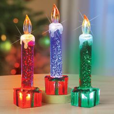 three brightly colored candles sitting next to each other on top of a table with christmas lights in the background