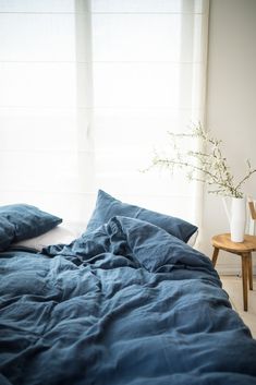 an unmade bed with blue sheets and pillows in front of a white curtained window