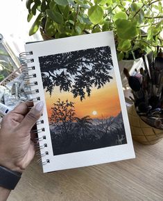 a person holding up an open notebook with a painting on the cover and trees in the background