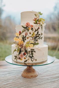 a wedding cake with flowers on it sitting on top of a wooden table in front of a grassy field